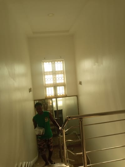 A young man wearing a green sleeveless shirt and camouflage shorts stands on a staircase, holding a white object. He is on the right side of the image, partially obscured by a metal railing. The staircase walls are painted white, and there is a window with a decorative metal grill on the upper part of the wall. A large mirror is visible on the wall below the window. The lighting suggests an indoor environment, likely within a building in Hotoro North, Nigeria, given the context.