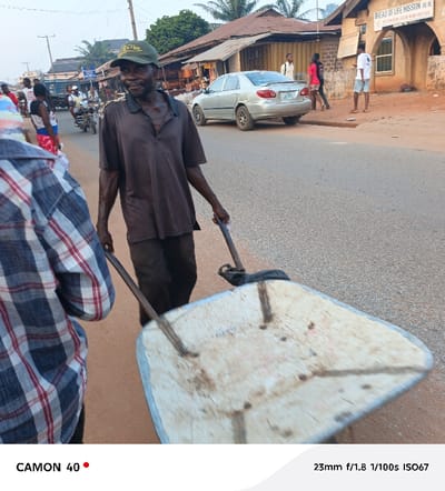 An older man in a brown shirt and green cap pulls a white wheelbarrow down a paved road. Other people walk along the street and a silver car is parked nearby. A building labeled "Bread of Life Mission" is in the background. The image is taken from the perspective of someone standing near the wheelbarrow. The photo's details indicate it was taken with a CAMON 40 camera at 23mm, f/1.8, 1/100s, and ISO67.