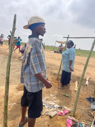 The image shows an outdoor scene, potentially in Maiyanga, Nigeria, with a dirt/sandy ground and sparse vegetation in the background. In the foreground, there are vertical bamboo-like poles suggesting a structure or makeshift shelter. A young person wearing a backward baseball cap, a blue and gray plaid shirt, and dark shorts stands facing away from the camera. An older woman in a light blue top and dark skirt stands further back. Scattered on the ground are personal items like flip-flops, clothing, and broom. There are other people visible in the middle distance, appearing to be carrying items. The sky is overcast.