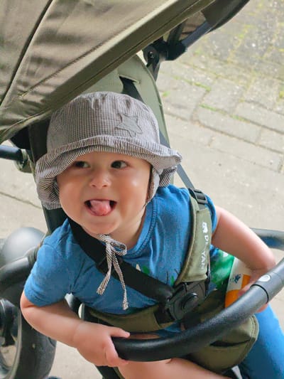 A baby wearing a grey and white checkered hat with a star emblem, a blue t-shirt, and a green and black safety harness is sitting in a stroller. The baby is sticking its tongue out and looking to the left. The stroller is outdoors, with a grey paved ground visible in the background. The stroller's canopy is green and appears to be made of fabric. The image captures the baby's face and upper body, as well as parts of the stroller and the surrounding environment. The specific location within the city of Georgsmarienhütte, Germany, cannot be determined from the provided image.