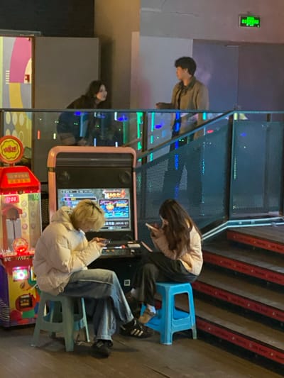 Three people are near arcade games. One person stands behind the arcade machine while two people sit. One seated person wears a cream-colored puffer jacket and blue jeans, looking at their phone. The other wears a tan top with dark bottoms and has long hair. There are stairs with red and black stripes in the background. The arcade games are red, pink, yellow, and purple.