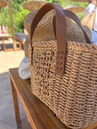 This is a close-up, eye-level shot of a woven straw bag with leather handles, resting on a wooden table. The bag is the primary subject and is in focus. The background is softly blurred, showing hints of beach umbrellas and lush greenery, suggesting an outdoor setting, possibly a resort or beachside cafe in Chernomorets, Bulgaria. No people are clearly visible, but there are faint suggestions of figures in the distant background. The lighting is natural and bright, indicative of daytime.