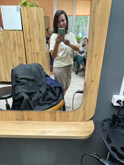 The image depicts a woman taking a selfie in a salon. She is holding a turquoise smartphone and wearing a white t-shirt and light-colored shorts. In the background, another woman is visible sitting in a chair, looking to the side. A chair covered with a black cloth is also present. The salon environment features wooden cabinets, a large mirror with a wooden frame, and a shelf below the mirror. The wall is painted a dark gray. A hair styling tool holder with brushes and what appears to be a hairdryer is visible on the right side of the image. The scene is consistent with the interior of a beauty salon. The location is specified as Kumyen, Russia.