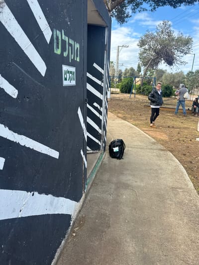 The image depicts an outdoor scene in Beit Yitshak Sha'ar Hefer, Israel. A concrete shelter structure occupies the left foreground, painted black with prominent white diagonal stripes. The Hebrew word "מקל" (Miklát), meaning 'shelter,' is painted in green on its wall, with a smaller white sign below it also displaying the word. An open doorway is visible on the right side of the structure. A black backpack rests on the concrete ground directly in front of this entrance.
To the right, the environment transitions to a dirt and dry grassy area, scattered with pine cones and sparse vegetation. A woman, wearing a black puffer jacket, white hoodie, black leggings, and brown shoes, stands in this area, smiling and looking towards the camera. Further into the background, a man is visible holding a child, with another individual partially obscured, appearing to be seated or crouching. Playground swings are discernible in the mid-distance. The background also features several trees, including a distinctly leaning tree, utility poles with power lines, and a partly cloudy sky.