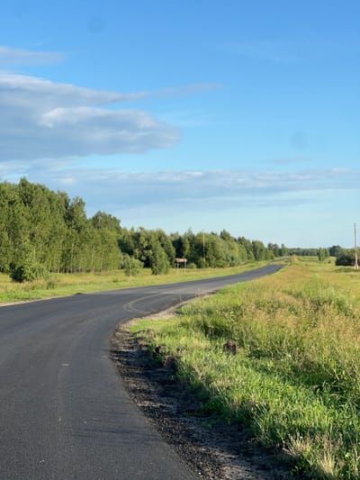 The image shows a paved road curving through a rural landscape under a bright blue sky with scattered white clouds. The road is dark asphalt, appearing freshly laid in some sections with visible tire marks. To the right of the road is a dense field of tall green grass, with some patches of dry, lighter-colored vegetation. Further to the right and stretching into the distance is a line of trees, predominantly deciduous, with lush green foliage.
On the left side of the road, there is a line of trees and bushes, also appearing green and healthy. In the distance, power lines and utility poles are visible, suggesting the presence of infrastructure. The overall scene depicts a scenic, possibly remote, rural setting. The lighting suggests it is daytime, likely late morning or afternoon, with bright sunlight casting shadows. The weather appears to be fair and warm. There are no people or animals visible in the image, nor any discernible text. The setting is characteristic of a rural road in a temperate climate.