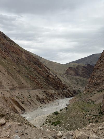 A wide-angle, eye-level shot shows a view of a river flowing through a mountainous valley under a cloudy sky. The river, appearing as a grey, agitated ribbon, snakes through the center of the frame, flanked by steep, rocky slopes. The mountainsides are a mix of barren, brown earth and scattered green vegetation, with visible layers of rock formations creating a textured, rugged landscape. A narrow, winding road is visible on the left slope, disappearing into the distance. Power lines are strung across the upper portion of the image, hinting at distant human presence. The overall impression is one of a remote and wild natural environment. The image is not specific to Reru, India, but rather depicts a generic mountainous river valley.