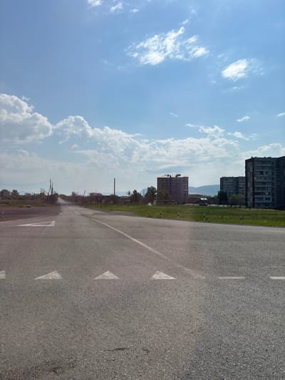 The image depicts a road in Chernogorsk, Russia. The road is paved and has lane markings, including directional arrows. On either side of the road, there is a grassy verge. In the background, there are several multi-story apartment buildings and what appears to be industrial structures. The sky is blue with scattered white clouds. There are no people visible in the image. The scene appears to be a typical urban or semi-urban landscape.
