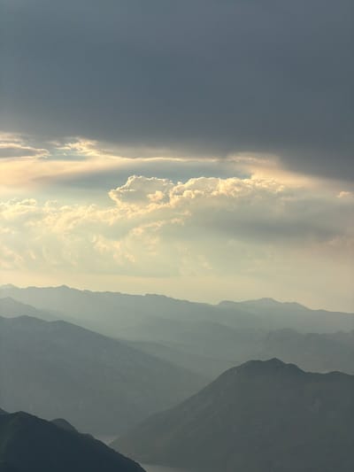 The image shows a dramatic sky filled with dark storm clouds above layers of misty mountains. The clouds are illuminated from behind by the sun, creating a glowing effect around the edges and highlighting some fluffy white clouds in the middle ground. The mountains recede into the distance, their peaks becoming increasingly obscured by atmospheric haze, creating a sense of depth and scale. The scene suggests a wild, untamed natural landscape, likely in a mountainous region. Based on the visual cues of the mountainous terrain and potentially coastal features, this image could be of the area around Kuk, Montenegro, a town located on the Bay of Kotor. The photograph captures a moment of changing weather, with the impending storm casting a moody atmosphere over the majestic landscape. There are no people, animals, or man-made structures prominently visible in this expansive natural vista.