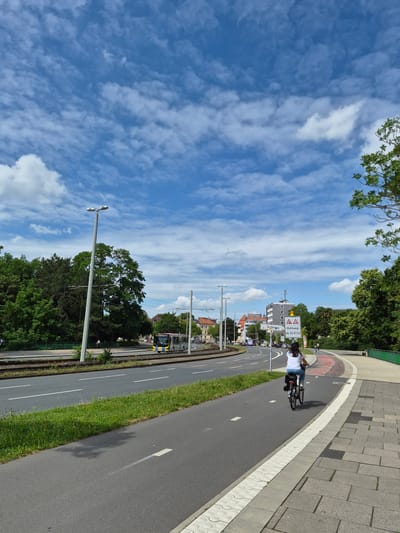 In Brunswick, Germany, a cyclist on a bicycle rides down a dedicated bike path next to a road with a tram line. The cyclist, a person with dark hair wearing a white t-shirt and blue jeans, is positioned in the foreground, riding away from the camera. The bike path is paved and has white lane markings. To the left of the bike path is a grassy area and then a road with traffic and a tram. A tram is visible in the distance on the tram tracks. Beyond the road, there are trees and buildings. The sky above is blue with scattered white clouds. A street sign is visible on the right side of the road, indicating "Schloss" and "Bohlweg ab 02.07.26". The overall scene depicts a sunny day in an urban environment with infrastructure for cycling and public transportation.
