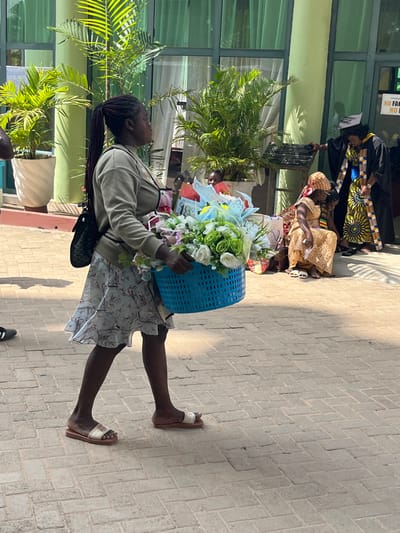 This image depicts an outdoor scene in Madina, Ghana, characterized by bright daylight. In the foreground, a dark-skinned woman with long braided hair pulled into a ponytail walks from left to right across a paved surface. She wears a light-colored long-sleeved top, a patterned knee-length ruffled skirt, and open-toed slide sandals, carrying a black crossbody bag on her left shoulder. She holds a large, light blue perforated plastic basket filled with artificial flowers, including white, green, light pink, and light blue blooms and ribbons.
The ground is composed of light-colored rectangular interlocking pavers arranged in a herringbone pattern. In the background, a modern building with large green-tinted glass windows and light green structural pillars is visible. Several potted plants, primarily palms, are positioned along the building's facade. To the right, a person in a black graduation gown with yellow and blue patterned trim and a black mortarboard cap stands near the building. Below them, two individuals are seated on a low step or the ground; one is clearly visible in a gold-patterned traditional dress. A white sign with partial red text, possibly reading "NO FAC...", is affixed to a pillar of the building behind the figures on the right. Shadows suggest a strong overhead light source.