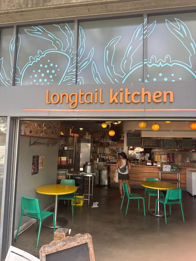 The image shows the exterior of a restaurant called "Longtail Kitchen" in New Westminster, Canada. The restaurant has a gray facade with large windows featuring light blue crab silhouettes. Orange lettering spells out the restaurant's name above the entrance. The entrance is open, revealing the interior of the restaurant. Inside, there are shelves with food items, a counter where two employees are visible, and orange paper lanterns hanging from the ceiling. In front of the restaurant, there are two small tables with yellow tops and green chairs. A woman wearing a white tank top and camouflage shorts is standing in front of the counter, facing away from the camera. The overall environment appears to be an urban setting during the daytime.