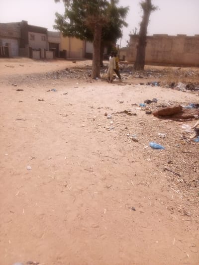 The image depicts an outdoor scene in Tudun Murtala, Nigeria, characterized by a predominantly reddish-brown, sandy dirt ground covering the foreground and mid-ground. The ground is uneven and littered with various pieces of refuse, including blue and white plastic bags, wrappers, and dried organic debris. In the mid-ground, two individuals are present near two prominent trees with visible trunks and sparse green and dry foliage. One individual wears a long, light-colored, possibly white or cream, garment and stands slightly ahead of the second individual. The second individual wears a darker, possibly yellow or ochre, garment. Both individuals appear to be walking or standing, facing generally towards the right side of the frame. In the background, multiple single and multi-story buildings with various architectural styles are visible on the left, alongside a tall, light-colored wall extending across the right. The sky is hazy and bright, indicating daylight conditions.