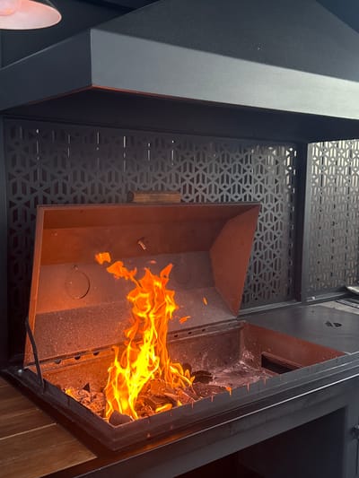 The image shows a close-up view of a large, modern outdoor grill with a fire burning inside. The grill is made of dark metal and has a hinged lid that is open, revealing a fire pit filled with glowing embers and flames. The background features a decorative metal screen with a geometric pattern. The overall scene suggests an outdoor cooking area, possibly a patio or backyard. The location is identified as Starokorsunskaya, Russia.