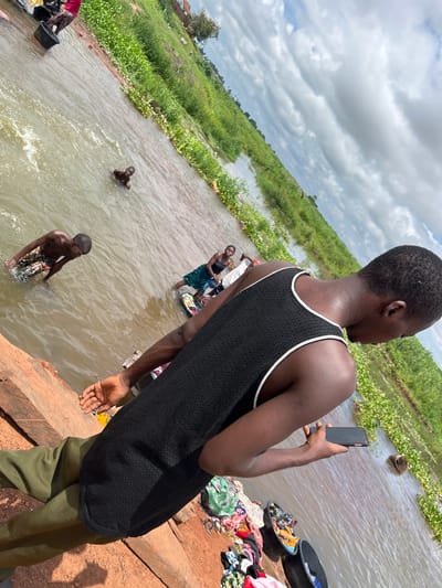 The image depicts a scene in Idah, Nigeria, centered around a body of water, likely a river or stream, under a cloudy daytime sky. In the foreground, a person is seen from behind and slightly to the right, wearing a black mesh tank top with white trim and olive green pants. This individual holds a black smartphone in their right hand, looking out towards the water.
To the right of this person, on the reddish-brown earth bank, are piles of colorful clothing and two dark wash basins, one containing clothes. The water itself is rippled and lined with dense green vegetation, both on the banks and floating on the surface in some areas.
Several other individuals are present in or near the water. Further into the water, a person wearing a patterned garment is partially submerged, appearing to be washing or bathing. Further back and to the left, another individual stands in the water, seemingly engaged in laundry with a basin. A third, smaller figure is seen partially submerged further away in the water. The background consists of lush green growth along the far bank and a wide expanse of grey, cloudy sky. No text is visible in the image.