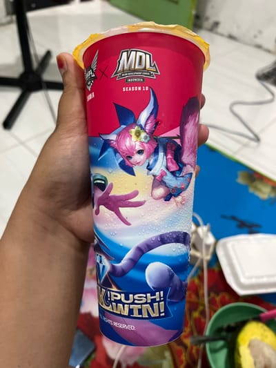 A hand holds a disposable cup with a yellow plastic lid. The cup is predominantly red with an illustration of a cartoon character with pink hair and a blue and pink outfit. The character appears to be flying or leaping. Text on the cup reads "MDL", "MLBB DEVELOPMENT LEAGUE", "INDONESIA", "SEASON 10", and "PUSH! WIN!". The background is blurry but suggests an indoor setting with some objects that appear to be part of a table and floor. There is no information in the image to indicate the location is the city of Cadek, Indonesia.