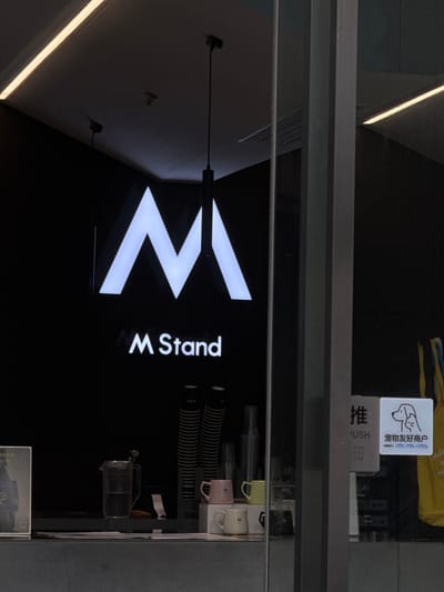 The image shows the interior of a business called "M Stand," likely a cafe, located in Xiangzhou, China. A large, illuminated "M" logo is prominently displayed on a black wall. Below the logo, the text "M Stand" is also illuminated. In the foreground, on a counter, are various items including cups, a french press with water, and stacks of paper cups. To the right, there is a glass door with a sign that says "PUSH" and features an icon of a dog and a cat, with Chinese text below it, indicating it is a "Pet Friendly Merchant." Yellow and blue fabric is visible to the far right. The overall lighting is dim with bright accents from the signage and ceiling lights.