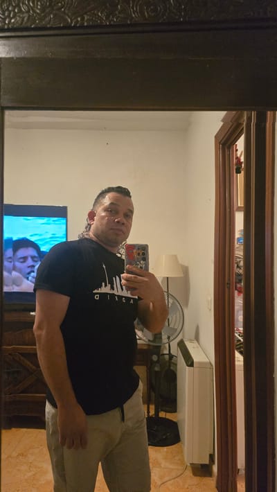 The image, captured from a mirror's reflection and originally oriented sideways, shows a man taking a selfie indoors. The man, who appears to be in his 30s or 40s, has short dark hair, stubble, and is looking directly at the camera with a neutral expression. He is wearing a black t-shirt with a white skyline graphic and the text "ADRID" (likely part of "MADRID") printed on it, along with light-colored pants. He holds a smartphone with a colorful case featuring a partial Mickey Mouse design. He is also wearing earrings.
The room reflected in the mirror has a tiled floor, appearing to be light brown or tan. The walls are light-colored, possibly white or off-white. On the left side of the reflection, a television screen displays what looks like a person's face and some blue, watery imagery. Below the TV, a dark wooden furniture piece is partially visible. On the right side of the reflection, a white table lamp with a rectangular shade and a white oscillating fan are present. Further back, a larger white appliance, possibly an air conditioner, is mounted higher on the wall. The mirror itself has a dark frame, likely wood, with decorative carvings on the visible left edge. The lighting suggests an indoor setting, possibly during the evening or night.