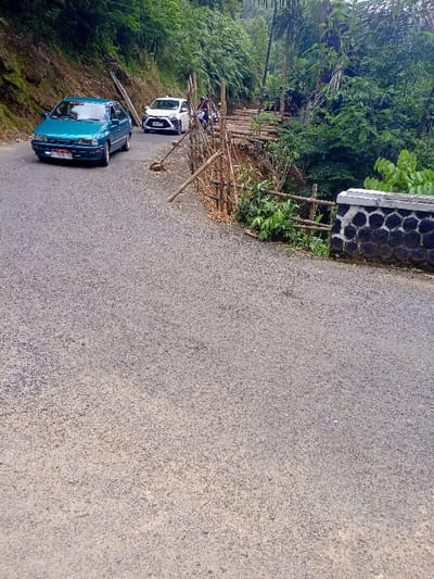 The image shows a paved road in Cisewu, Indonesia (latitude: -7.41, longitude: 107.542). A teal car is driving away from the camera. There is another white car parked in the background. The road is flanked by dense vegetation on both sides, with a section of damaged road on the right side. There is a short section of a stone wall on the right side of the image. The wall is built of black and white stones and has a section of wooden railing in front of it.