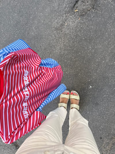 A person stands on an asphalt surface, wearing white pants and light-colored sandals. The person is holding a folded garment that has alternating red and white vertical stripes on one side and blue vertical stripes on the other. The asphalt surface is grey and appears to be a street or sidewalk, with some small pebbles and a crack containing a discarded cigarette butt visible. The image is taken from a top-down perspective, looking down at the person's feet and the garment. The location is not specified in the image, nor does it provide any identifiable landmarks for Stockholm, Sweden.