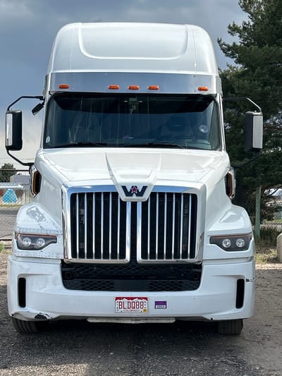 A white semi-truck is parked on a gravel lot. The truck is facing the camera and is centered in the frame. The truck has a large chrome grille and the Western Star logo in the center. The truck's license plate reads "BLDO88 COLORADO". The sky above is overcast and there are trees in the background to the right of the truck. The location is the city of Aurora, United States of America.