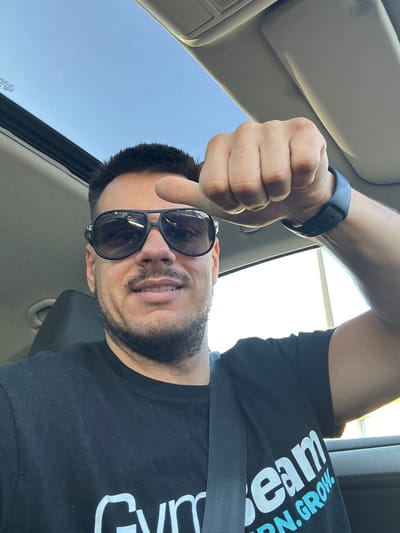A man, wearing a black t-shirt with white and blue lettering that reads "Gym Team", sunglasses, and a blue wristband, is inside a vehicle. He is looking at the camera and giving a thumbs-up gesture with his right hand. The car's sunroof is open, revealing a clear blue sky. The photo appears to have been taken in Busmantsi, Bulgaria, judging by the license plate visible through the sunroof.
