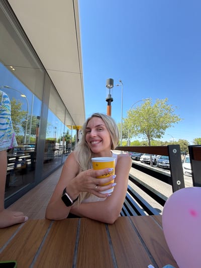 A woman is holding a yellow cup and smiling at the camera. She has blonde hair and is wearing a white top. She is sitting at a wooden table outdoors, with a building with large windows to her left and a street with cars to her right. A pink balloon is visible in the lower right corner of the image. The location is Húmera-somosaguas-prado Del Rey, Spain.