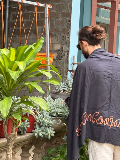 In Mai Chau, Vietnam, a person with a man bun and sunglasses stands facing away from the camera, wearing a dark grey garment with orange text on the back. They are holding a mobile phone in their left hand. The scene features several potted plants, including a tall plant with broad green and yellow leaves in a red pot, and a collection of succulents in grey pots. A rustic balcony railing and a brick wall are visible in the background, along with a clothesline with orange ropes and a metal structure. The overall environment suggests an outdoor setting in a residential area.