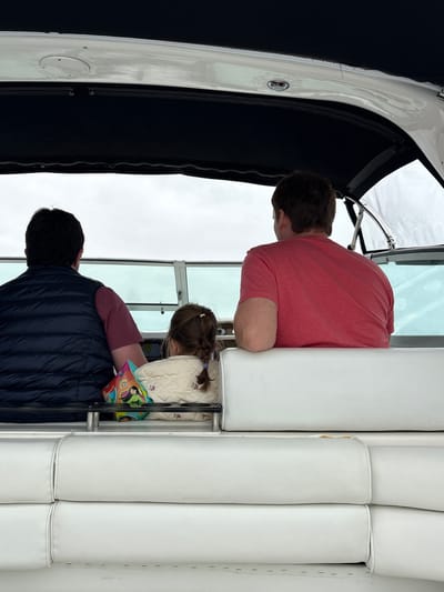 Three people are seated on a boat. The subject in the foreground is a man wearing a dark blue puffer vest over a maroon shirt. He is facing away from the camera, looking towards the right. Behind him and to his right, a young girl with braided brown hair is seated. She is wearing a quilted cream-colored jacket and is holding a colorful object with cartoon characters. She is also facing away from the camera, looking downwards. In the background and to the left, a man in a coral-colored t-shirt is seated, facing away from the camera. The boat has white seating and a black canopy. The overall scene appears to be a daytime activity on the water, possibly a scenic tour. The weather appears overcast.