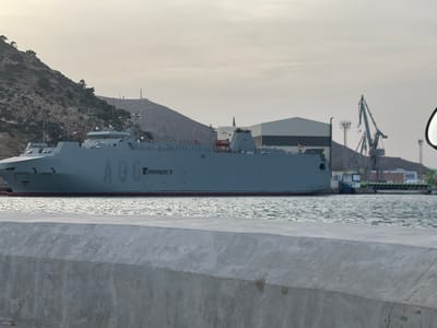 A large, gray naval vessel with the markings "A96" on its side is docked in the port of Cartagena, Spain. The ship is positioned in the middle ground, with a rocky, tree-covered hill rising behind it on the left side of the frame. In the background, there are industrial buildings, cranes, and antennas, indicative of a port or naval facility. The water in the foreground is choppy and reflects the overcast sky. A concrete barrier runs along the bottom of the frame, suggesting the viewer is on a quay or a similar elevated platform. The lighting suggests it is daytime, possibly late afternoon or early morning due to the soft, diffused light and muted colors.