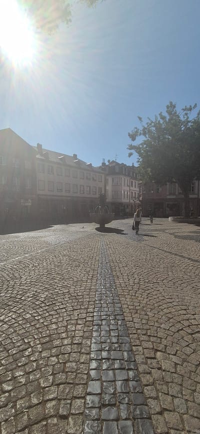 This is a vertical, medium-shot, outdoor photograph taken on a sunny day in the city of Mainz, Germany. The image depicts a cobblestone plaza with a fountain and several buildings in the background.
The foreground is dominated by the cobblestone ground, which is laid in concentric circles. A dark, reflective strip runs down the center of the plaza, appearing to be made of polished stone or metal.
In the middle ground, a woman with long, light-colored hair walks away from the camera, wearing a white top and dark pants, carrying a dark bag. To her left, a fountain with a sculpture is visible. Further in the background, several people are walking or standing.
The background features a row of traditional European buildings with multiple windows and pitched roofs. A large tree with dense foliage is on the right side of the frame. The sky is clear and blue, with the sun creating lens flare in the upper left corner. The overall impression is of a tranquil, sunny day in a European town square.
