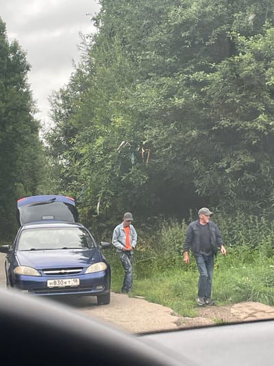 In Gavrilovka, Russia, a blue Chevrolet sedan with its trunk open is parked on the side of a dirt road. Two men are standing near the vehicle. The man on the left, wearing a light blue denim jacket over an orange shirt and blue jeans, stands by the car with his hands clasped in front of him. The man on the right, dressed in a dark jacket, blue jeans, and a grey baseball cap, stands a few feet away, looking to his left. They are surrounded by lush green foliage, with trees and tall grass forming the backdrop. The sky is overcast, suggesting a cloudy day. A cluster of colorful ribbons are tied to the branches of a nearby tree. The license plate of the car is visible and reads "H830KT 18".