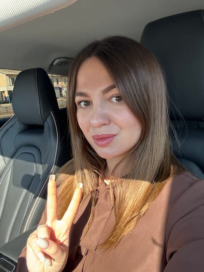 A female subject is depicted in a close-up, chest-up shot taken inside a vehicle, identified as being in Likino-Dulyovo, Russia. The subject, appearing to be in her 20s or 30s, has medium-length brown hair, brown eyes, and a light complexion, with light pink lipstick on her lips. She is wearing a brown long-sleeved top. Her right hand is raised, making a "peace" sign (V-sign) with the index and middle fingers extended. Her fingernails are square-shaped with light-colored polish, and a silver ring is visible on her right ring finger. She maintains direct eye contact with the camera and exhibits a faint, closed-mouth smile. The interior of the vehicle features black leather seating with light-colored stitching, visible on the headrest and seat to her left. Sunlight illuminates the scene, casting shadows across her hair and right arm. Through the vehicle's window, behind the car's headrest, several light-colored, multi-story residential buildings are visible, one of which appears unfinished or in a state of disrepair. The ground outside appears unpaved.