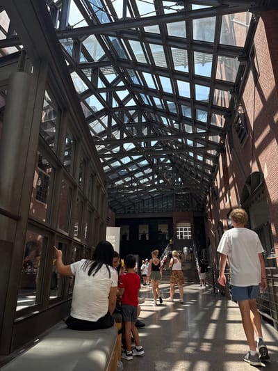 The image shows an indoor scene at the National Museum of American History in Washington, D.C. The architecture features a large, angular glass and steel atrium ceiling, with sunlight filtering through the panes and casting dappled shadows on the floor. Brick walls with multiple windows line the sides of the atrium.
In the foreground, a person with long dark hair, wearing a white t-shirt and black shorts, is sitting on a padded bench, gesturing with their right hand. To their right, a young boy in a red t-shirt and gray shorts stands next to them. Further down the walkway, a young person with curly blond hair, wearing a white t-shirt and blue shorts, walks away from the camera. Other museum visitors are visible walking up a grand staircase in the background. A banner with text is also visible on the left side of the image. The overall atmosphere appears to be a busy day at a museum.