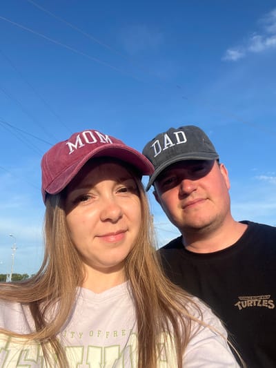 A young couple is taking a selfie outdoors under a clear blue sky. The woman is on the left, wearing a maroon baseball cap with "MOM" embroidered in white. She has long, blonde hair and is wearing a light purple t-shirt. The man is on the right, wearing a dark baseball cap with "DAD" embroidered in white. He has short brown hair and is wearing a black t-shirt with the Teenage Mutant Ninja Turtles logo on it. They are standing close together, with the woman's head tilted slightly towards the camera and the man looking directly at the camera. The background shows a few power lines stretching across the sky, and some distant trees and streetlights. The image was taken in Mal'tsevo, Russia.