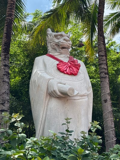 A stone statue of a mythical creature with a dragon-like head stands outdoors, adorned with a large red silk ribbon tied in a bow around its neck. The statue is positioned between two palm trees and amidst lush green foliage. The creature's head is detailed with sharp teeth and an open mouth, and it wears traditional robes. In its right hand, the statue holds a rolled scroll. The background consists of dense green trees and a bright, slightly cloudy sky. The scene appears to be in Xinzheng, China.