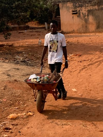 An image taken in Otuocha, Nigeria, depicts a young Black male pushing a single-wheeled, rusty metal wheelbarrow filled with assorted refuse. The male, positioned centrally, wears a white t-shirt with black trim and a graphic print, dark trousers, and dark open-toed sandals. Multiple beaded necklaces and pendants are visible around his neck, along with a silver-colored watch on his left wrist. He looks directly at the viewer with a neutral expression, gripping the wheelbarrow's handles.
The wheelbarrow contains various discarded items including crushed aluminum cans, plastic containers, pieces of wood, and a large, dark organic mass resembling an animal carcass or root. The scene unfolds on an unpaved, reddish-brown dirt path, characteristic of an undeveloped or semi-rural area. In the background to the right, a weathered, light brown block building with a small rectangular opening is visible. Sparse green vegetation and trees are present in the background to the left. A white bird, possibly a chicken, is visible on the ground in the middle distance near the building. The ground in the foreground shows scattered light-colored debris. The area is illuminated by bright daylight, casting short shadows.