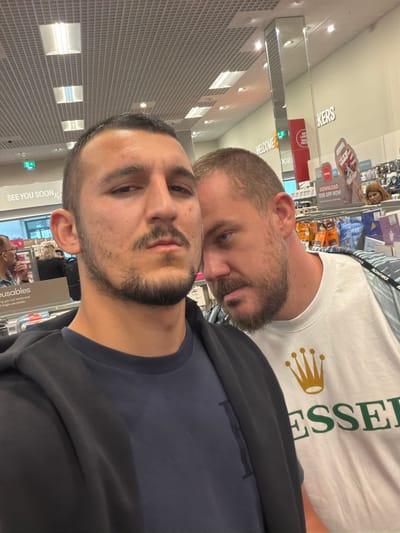 Two men are captured in a close-up, likely a selfie, within an indoor retail environment in Luton, United Kingdom. The image is taken from a slightly high angle, looking down.
In the foreground, a man with dark hair, a beard, and a mustache looks directly towards the camera with a neutral expression. He is wearing a dark blue t-shirt under a black jacket or hoodie.
Behind him and slightly to his left, another man with a beard is visible. He is wearing a white t-shirt with a gold crown logo and the green text "ESSER" (likely part of "ESSENTIAL" or similar) printed on it. He has a lanyard around his neck with "MONTIREX" printed on it. This man is looking downwards and slightly to the left of the camera.
The background shows typical elements of a retail store, with shelves displaying various products, though specific items are not clearly identifiable. Bright overhead artificial lighting illuminates the scene. Several signs are visible in the background. On the wall or a display, white text reads "SEE YOU SOON X" and "WELCOME." Other signs include "DOWNLOAD OUR APP NOW," "BIG LABEL SMALL PRICE," and "KERS" (possibly part of a word like "Bakers" or "Snackers"). Further text mentioning "reusables" and "cut waste and single use plastic" is also discernable on a sign. A woman with light brown hair is partially visible behind the second man, further in the background.