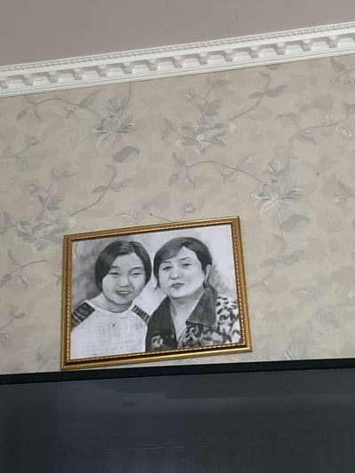 A framed black and white portrait of two women hangs on a wall with floral wallpaper. The woman on the left, appearing younger, has short, dark hair and is smiling slightly. She wears a striped shirt. The woman on the right has dark hair pulled back and a neutral expression. She is wearing a patterned jacket. A white decorative molding runs along the top of the wall. The bottom of the image shows a dark, reflective surface, likely a television screen. The scene is indoors.