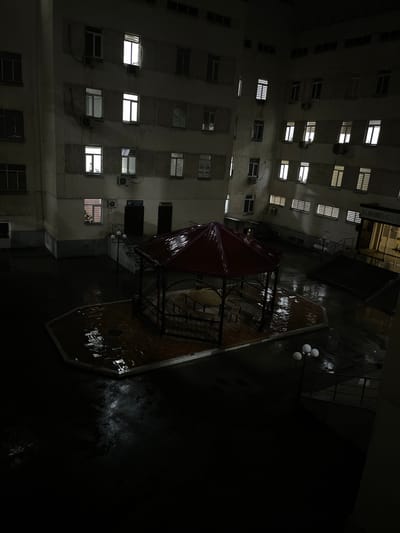The image, likely taken at night in Navoiy City, Uzbekistan, shows a courtyard surrounded by a multistory building. The courtyard is wet, possibly from recent rain, reflecting light. A red-roofed gazebo sits in the center. Three spherical light fixtures are visible near a shadowed area. The building facade has numerous windows, some illuminated. Air conditioning units are attached to the exterior walls below some windows. The perspective suggests the image was taken from an elevated position.
