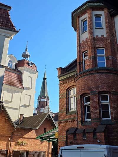 The image shows a street scene in Sopot, Poland, dominated by the architecture of several buildings. In the foreground, on the right, is a corner of a red brick building with white-framed windows and decorative green banding. To the left of this is another red brick building with a pitched roof and a sign reading "Lotto salon gier". Behind these, a tall, slender church spire with a green roof and white and red accents pierces the clear blue sky. To the far left, a cream-colored building with a red tiled roof and a small dome-like structure is partially visible. The scene is illuminated by bright sunlight, casting shadows and highlighting the textures of the buildings. There are no people clearly visible in the image. The overall impression is of an urban environment with historical architecture under a bright, clear sky.