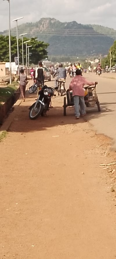 The media file depicts a street scene in Gboko, Nigeria. The foreground shows a dirt road with a parked black motorcycle on the left. To the right, a person in a pink shirt and blue jeans is pushing a two-wheeled cart. In the background, several people are walking or riding motorcycles, suggesting a moderately busy day. One person is wearing a striped shirt and appears to be on a motorcycle. Further in the distance, more people and vehicles are visible, and a mountainous landscape forms the backdrop under a cloudy sky. The scene appears to be taking place during daylight hours, with the lighting suggesting it might be late morning or afternoon. The environment appears somewhat dry and dusty. There is no visible text in the image.