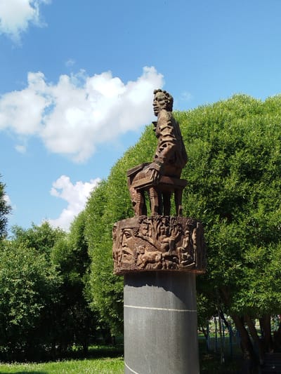 The photograph depicts a sculpture in Yalutorovsk, Russia, set against a backdrop of lush green trees and a bright blue sky with scattered white clouds. The sculpture is of a man seated on a stool, carved from a dark material, possibly bronze or wood. The man has distinct facial features and is looking to his left. The stool itself is supported by a cylindrical pedestal that is decorated with relief carvings of human figures and other motifs. The base of the sculpture appears to be a weathered, gray stone column. In the background, dense foliage from trees creates a vibrant green wall, and further back, a glimpse of a blue fence and some lampposts suggests a park or public space. The ground in the foreground is covered in green grass, with some white flowers or seed heads visible. The lighting suggests it is daytime, with sunlight casting shadows on the sculpture and illuminating the scene.