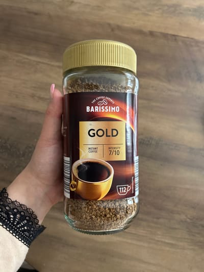 The image shows a close-up of a glass jar of Barissimo Gold instant coffee, held by a person's hand with manicured nails, against a wooden background. The label on the jar prominently displays "BARISSIMO GOLD" and "INSTANT COFFEE" with an intensity rating of "7/10". There is also an illustration of a cup of coffee and the number "112" with a coffee cup symbol. The lid of the jar is gold-colored. The person is wearing a light-colored garment with black lace trim on the sleeve. The wooden surface appears to be a table or countertop. No specific location in Waldbröl, Germany, is depicted or indicated in the image.