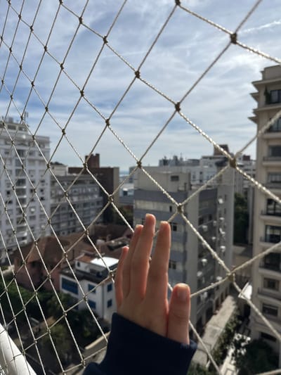 An elevated view of Porto Alegre, Brazil, is presented through a tightly-meshed, light-colored safety net. The grid of the net dominates the immediate foreground. In the lower-center, a human hand, appearing juvenile, with extended fingers, presses lightly against the net. A dark blue sleeve covers the forearm. Beyond the net, a dense urban landscape unfolds under a partly cloudy sky. Multi-story residential and commercial buildings of various heights and predominantly light hues populate the midground, interspersed with green foliage. The distant horizon reveals a body of water, consistent with the Guaíba. The scene is illuminated by natural daylight.