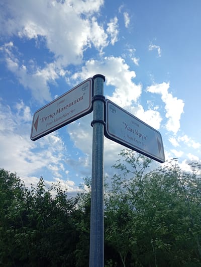 The image features a street signpost against a vibrant blue sky filled with scattered white clouds, indicating a bright, clear day. The photograph is taken from a low angle, looking up towards the signs and the sky, with green foliage, including trees and bushes with small white flowers, visible at the bottom and sides of the frame.

A metallic grey pole supports two rectangular street signs. Both signs have a white background with a red border and a small circular logo in their top-left corner.

The left sign, which points to the left, displays the street name in Cyrillic as "улица 'Петър Момчилов'" and its English transliteration "Petar Momchilov Str." below.

The right sign, pointing to the right, shows the street name in Cyrillic as "улица 'Хан Крум'" with its English transliteration "Han Krum Str." underneath.

The visible text on the signs indicates that the scene is in a Bulgarian-speaking region, specifically in Roza, Bulgaria, marking the directions for "Petar Momchilov Street" and "Han Krum Street." The overall setting suggests an outdoor, possibly residential or natural, environment.