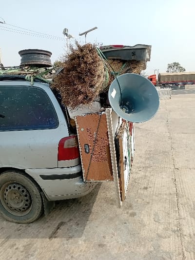 This image depicts a silver station wagon parked on a paved outdoor surface under a clear, bright sky in Kariya, Nigeria. The vehicle is overloaded with various objects. On its roof, a dark container or bundle is visible, alongside a large, irregularly shaped mass of dried, brown fibrous material, possibly brushwood or a collection of brooms, secured with green ropes. An old, dark-colored wheel rim also rests on the roof. Strapped vertically to the rear bumper are several flat panels, likely made of wood or composite material, extending outwards. A large, grey-blue horn-shaped loudspeaker is prominently attached to the right side of these rear panels. The vehicle's rear left tire and wheel show dirt accumulation, and the rear bumper exhibits minor scuffs. In the background, several large trucks, some with canvas-covered cargo areas, are parked. White barriers or fences are visible in the mid-ground, along with utility poles equipped with solar panels. The ground is a light-colored concrete or asphalt, showing some discoloration and texture. No people are visible in the scene.