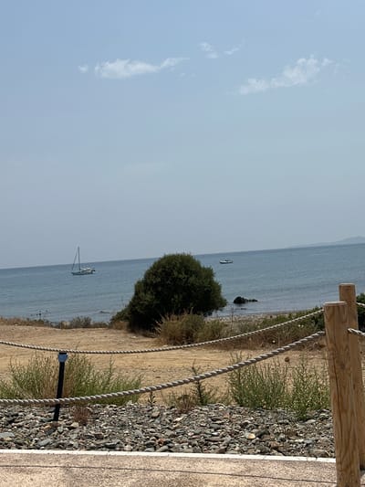 The image depicts a coastal scene under a clear, pale blue sky with wispy clouds. In the foreground, a gravel path is bordered by a wooden post and rope barrier. Beyond this, a sandy area with sparse, dry vegetation leads down to a pebble beach. The sea, a muted blue, stretches to the horizon. Two vessels are visible on the water: a sailboat with its mast and sails down, and a smaller boat with at least two individuals. A dense, dark green bush dominates the mid-ground on the left, with a few rocks visible near the shoreline. The distant coastline is hazy. The scene appears to be located in Biddeputzi/Villaputzu, Italy, based on the provided context.