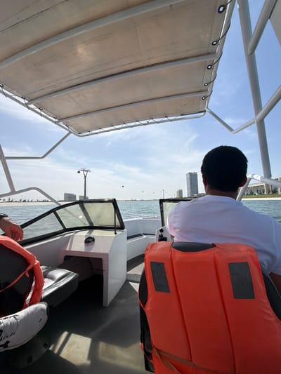 The image captures a first-person perspective from inside a boat, rotated 90 degrees clockwise. When viewed upright, a person with short dark hair, wearing a white t-shirt and an orange life vest, is seated in the foreground, facing away from the camera and towards the front of the boat. Their hands are out of view, suggesting they might be operating the boat or resting them on a control panel. Another person's feet, wearing light gray athletic shoes with a visible "On" logo, are visible in the lower right corner, resting on the boat's floor or a seat.
The boat features a white metal frame supporting a light-colored canvas canopy overhead, providing shade. A windshield with a black frame is visible towards the front of the boat, just beyond the seated person. The boat appears to be on a large body of water, likely a river or lake, with ripples visible on the surface.
In the background, under a clear, bright blue sky with sparse clouds, a distant shoreline is visible with several modern-looking buildings, suggesting a city or town. Stretching across the water in the mid-ground are prominent overhead cables, possibly part of a power line or a cable car system, with spherical insulators or gondolas suspended from them.
The scene depicts a sunny day, likely during late morning or early afternoon, with good weather for boating. The activity is a boat ride on the water, offering views of the urban landscape. The location context provided is Kuchtumgali, Uzbekistan.