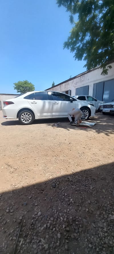 The image depicts an outdoor scene in Almalyk, Uzbekistan, with a focus on a white sedan. A person, dressed in light-colored pants and a grey top, is kneeling on the ground next to the front passenger side of the vehicle, appearing to be working on it. The car is parked on a gravel and dirt lot in front of a building with a white facade and large blue garage doors. The building has some Cyrillic text on its exterior wall. Other vehicles are partially visible in the background, including a light-colored sedan with a license plate starting with "06 J637 HA" on the right. The scene is under a clear blue sky with dappled sunlight casting shadows from nearby trees.
