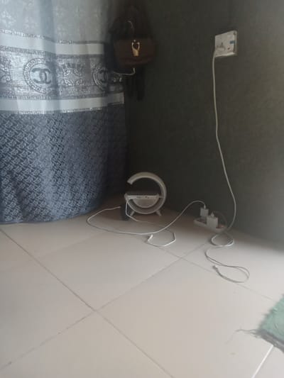 A corner of a room in a residential building in Zanzi, Nigeria, shows a tiled floor and a dark textured wall. To the left, a curtain with a repeating pattern of "CC" logos and diamond shapes hangs, partially obscuring a dark handbag with gold hardware suspended from a hook on the wall. In the center of the room, a white circular object with a silver base, possibly a speaker or lamp, rests on the floor with a white charging cable connected to it. A white extension cord with multiple plugs is situated near the wall, with two white adapters plugged into it, and a white cable originating from the extension cord trails across the floor. A white electrical outlet is visible on the wall to the right, with a white cable plugged into it.